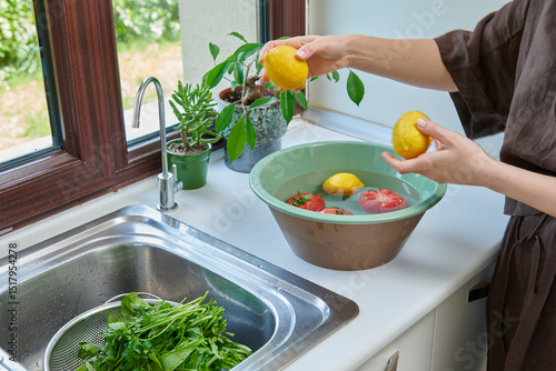 A woman washes fresh lemons, tomatoes, and leafy greens in a kitchen sink, preparing healthy ingredients near a sunlit window.