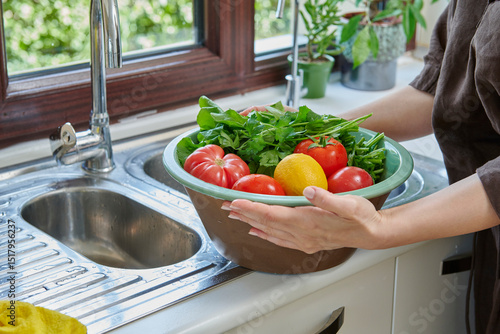 Photography A woman washes fresh lemons, tomatoes, and leafy greens in a kitchen sink, preparing healthy ingredients near a sunlit window