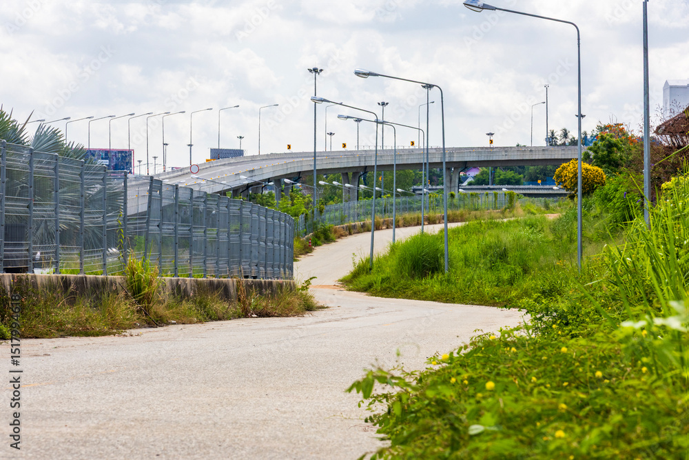 Urban elevated highway and small rural road with overgrown grass, street lights, and fence in foreground under bright blue sky. Transportation infrastructure in suburban area. Urban elevated highway and small rural road with overgrown grass, street lights, and fence in foreground under bright blue sky. Transportation infrastructure in suburban area.