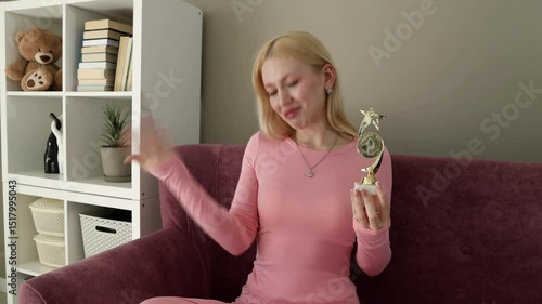 Blonde woman in pink loungewear sitting on a purple couch, joyfully holding a small golden trophy and gesturing as if giving a speech, with a bookshelf in the background
