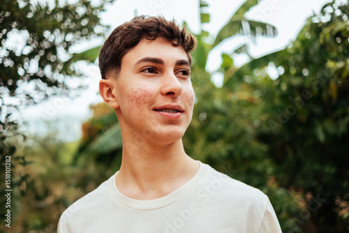Smiling teenage boy with acne and braces looking standing outdoors surrounded by green foliage