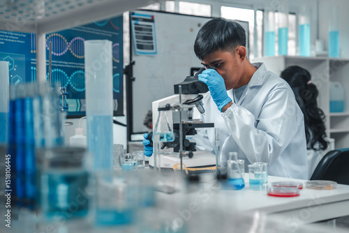 Focused Asian male scientist using microscope in modern laboratory with DNA structure chart in background. Concept of biotechnology, genetics research, molecular biology and advanced healthcare.