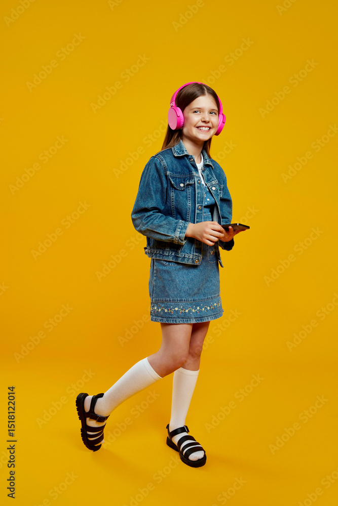 Fototapeta premium A smiling little girl wearing denim and pink headphones, holding a smartphone, enjoying music joyfully. Ideal for technology and lifestyle advertisements.