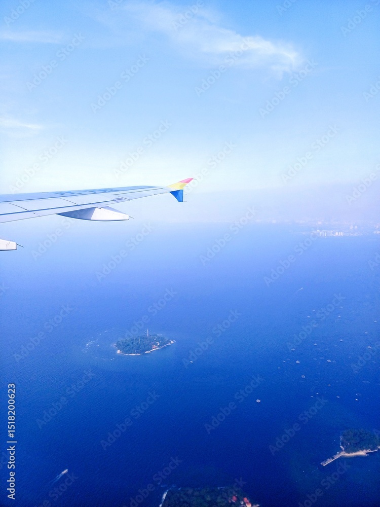 Naklejka premium Serene aerial view from an airplane, with a portion of the white aircraft wing visible. Below, the vast expanse of deep blue ocean stretches out, dotted with several lush green tropical islands