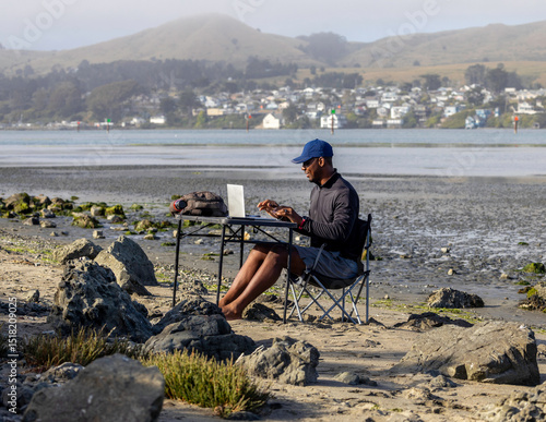 A man with a laptop working on a beach or bay at low tide.  Full-time RV nomad lifestyle, working in shorts from his home office.