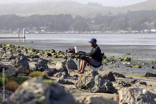 A man with a laptop working on a beach or bay at low tide.  Full-time RV nomad lifestyle, working in shorts from his home office.