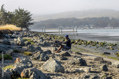 A man with a laptop working on a beach or bay at low tide.  Full-time RV nomad lifestyle, working in shorts from his home office.