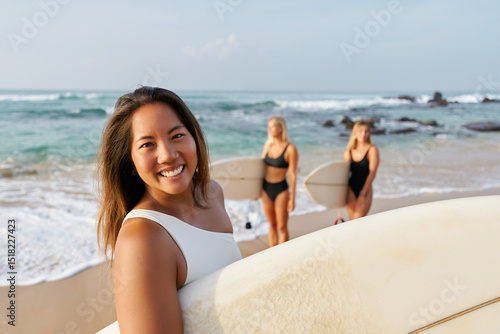 Wallpaper Mural Smiling woman on sandy beach holds surfboard. Friends in swimsuits behind prepare for surfing. Ocean waves and sunny weather. Summer fun, water sports, beach vibes. Torontodigital.ca