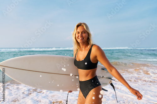 Photography Blonde woman in black bikini with surfboard on sandy beach