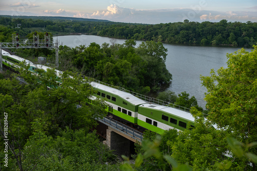 Aerial view of a train going through a lush green forest area by the lake in summer time at sunset hour. Commute, public transit, green transport, zero emission, travel, journey concept. 