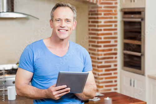 Картината върху платно Senior man holding tablet computer and smiling in modern kitchen at home with ga