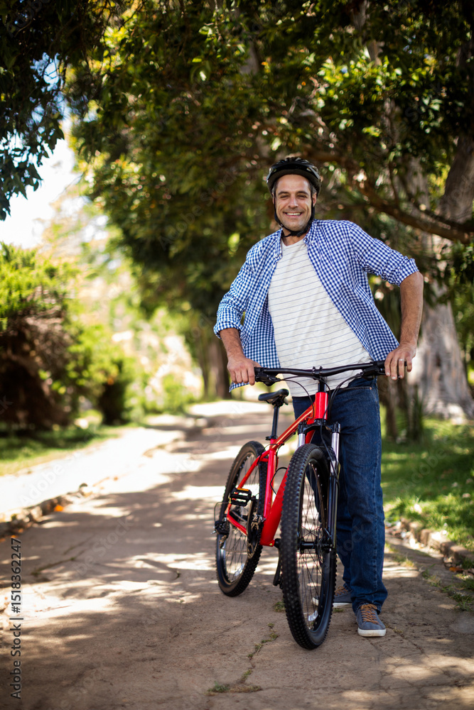 Naklejka premium Man standing holding red mountain bicycle on sunlit tree-lined path wearing cycling helmet