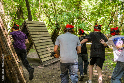 teenagers playing laser tag in pine forest, with guns, accessories, running between pine trees, back, wooden shelters, like huts, youth, childhood, entertainment, holiday, game, people