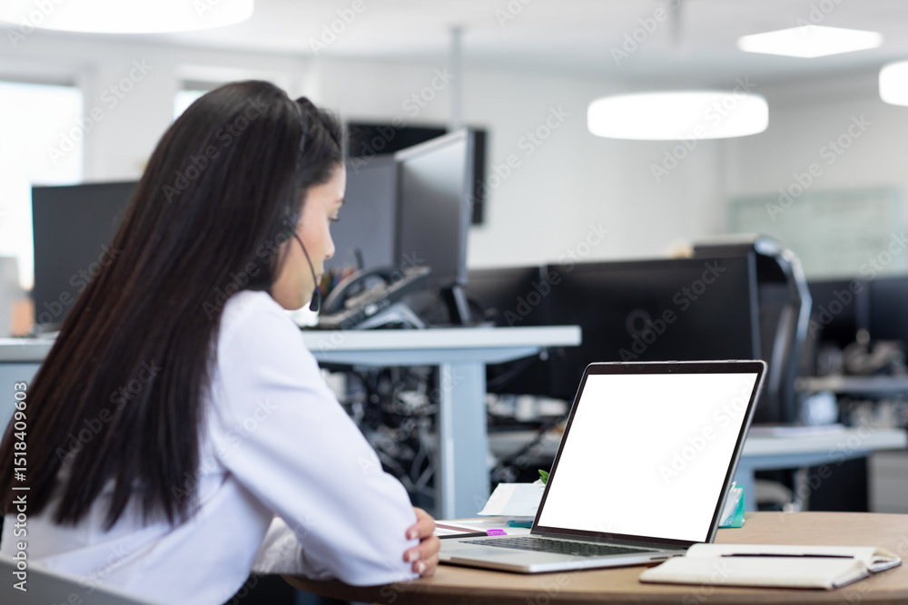 Office worker wearing headset typing on laptop at desk amid monitors in office with LED lights Office worker wearing headset typing on laptop at desk amid monitors in office with LED lights