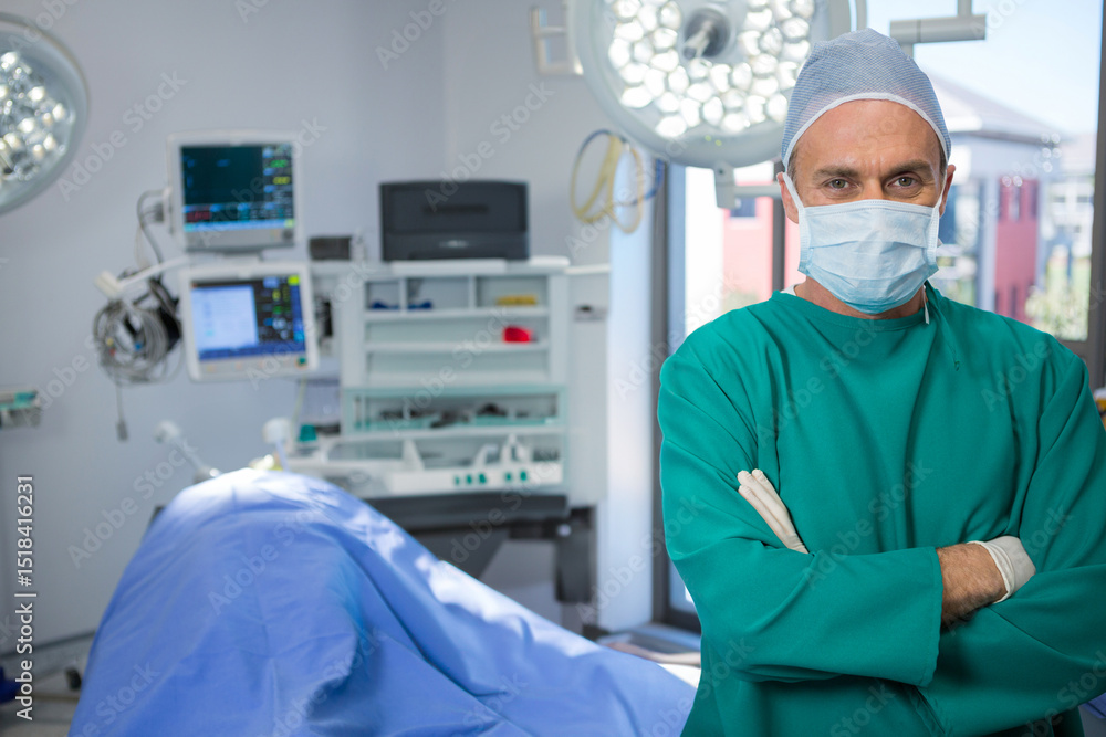 Male surgeon wearing scrubs and crossing arms in operating room under surgical lights, copy space Male surgeon wearing scrubs and crossing arms in operating room under surgical lights, copy space
