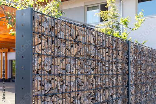 A modern gabion fence constructed from stones creates an elegant boundary. It stands in a contemporary setting, complemented by greenery and a sleek architectural backdrop, showcasing innovation