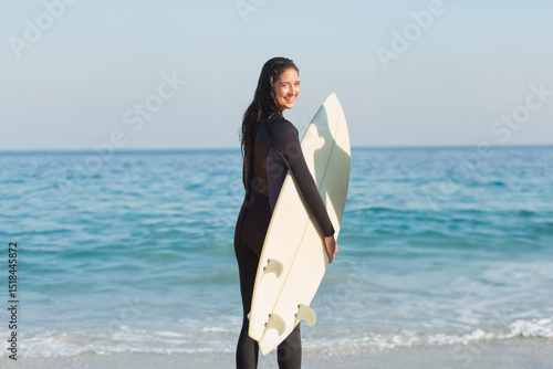 Female surfer wearing black wetsuit smiling while holding white surfboard on sandy beach