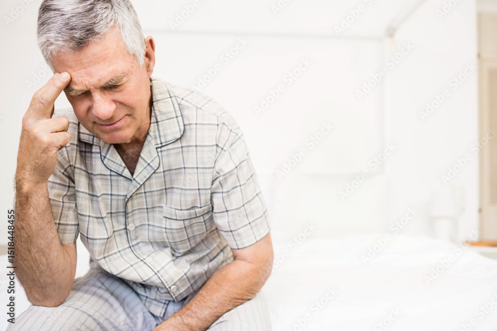 Fototapeta premium Senior man pressing temple while on bed edge in bedroom with lamp, plaid pajamas, copy space