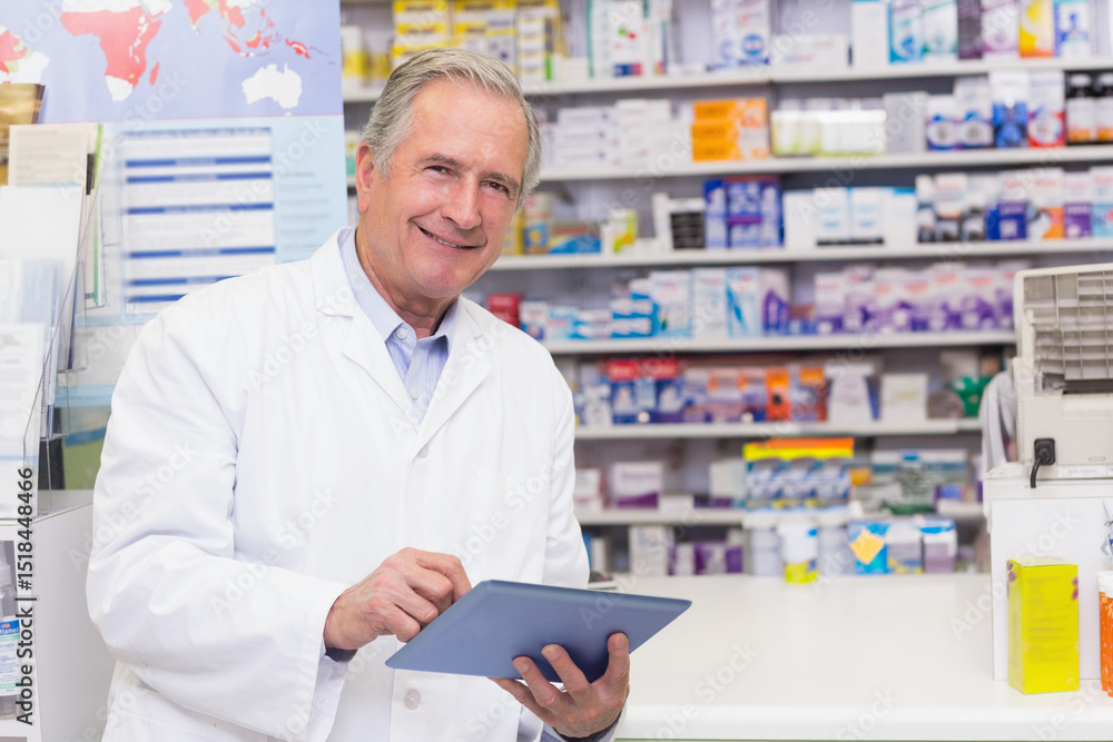 Senior male pharmacist using tablet behind pharmacy counter with brochure stand and medications Senior male pharmacist using tablet behind pharmacy counter with brochure stand and medications