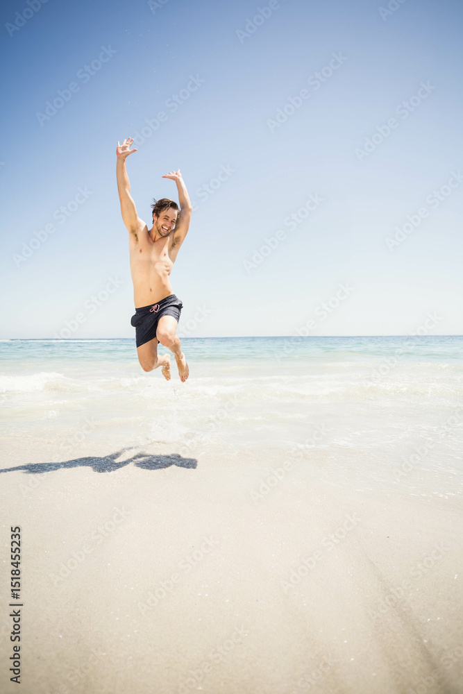Man jumping at sandy shoreline wearing dark swim trunks, splashing shallow water and casting shadow Man jumping at sandy shoreline wearing dark swim trunks, splashing shallow water and casting shadow