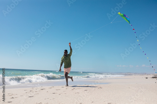 Running barefoot on beach, African American man wearing hoodie holding green blue kite, copy space