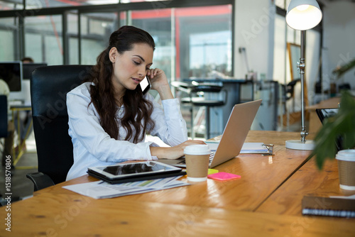 Woman talking on smartphone and typing on laptop in open-plan office with paperwork and coffee cups