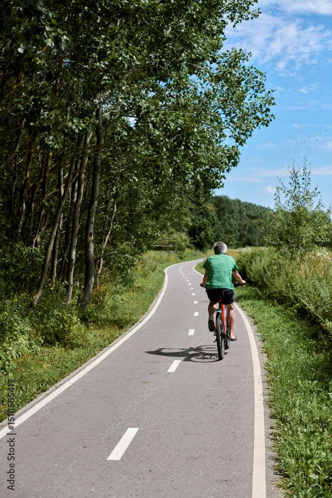 Fototapeta premium Elderly adult rides bicycle along serene park trail, surrounded by lush greenery under blue sky