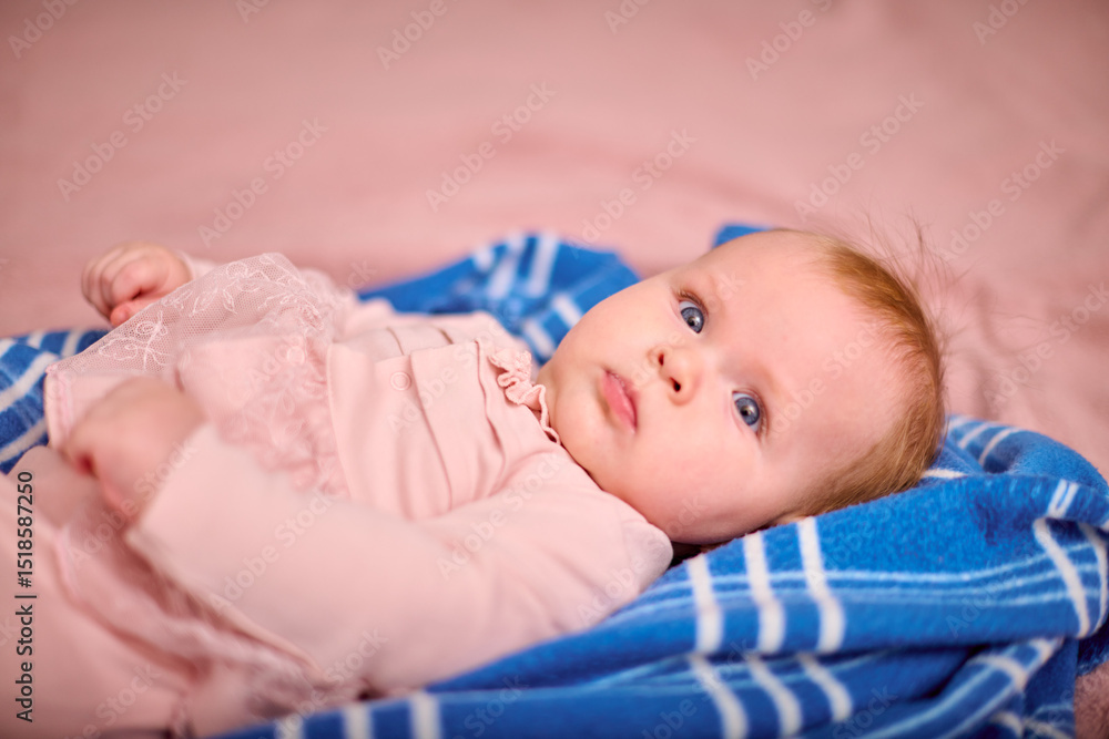 Infant female with red hair and fair skin lies on blue striped blanket. Soft lighting highlights pink outfit, creating serene and cozy setting Infant female with red hair and fair skin lies on blue striped blanket. Soft lighting highlights pink outfit, creating serene and cozy setting