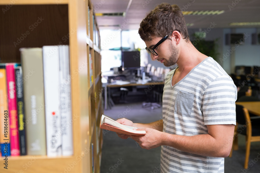 Man standing by bookshelf in library holding open paperback and reading next to computer monitors Man standing by bookshelf in library holding open paperback and reading next to computer monitors