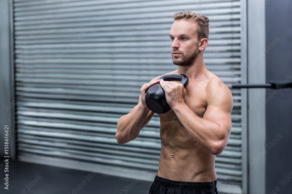 Obraz premium Shirtless adult male lifting kettlebell in gym area near metal shutter and barbell rack, copy space