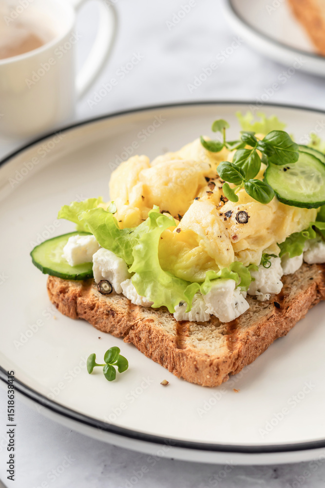 Scrambled eggs on grilled toast with salad leaves, cheese and micro greens on plate. Delicious breakfast or brunch close up Scrambled eggs on grilled toast with salad leaves, cheese and micro greens on plate. Delicious breakfast or brunch close up