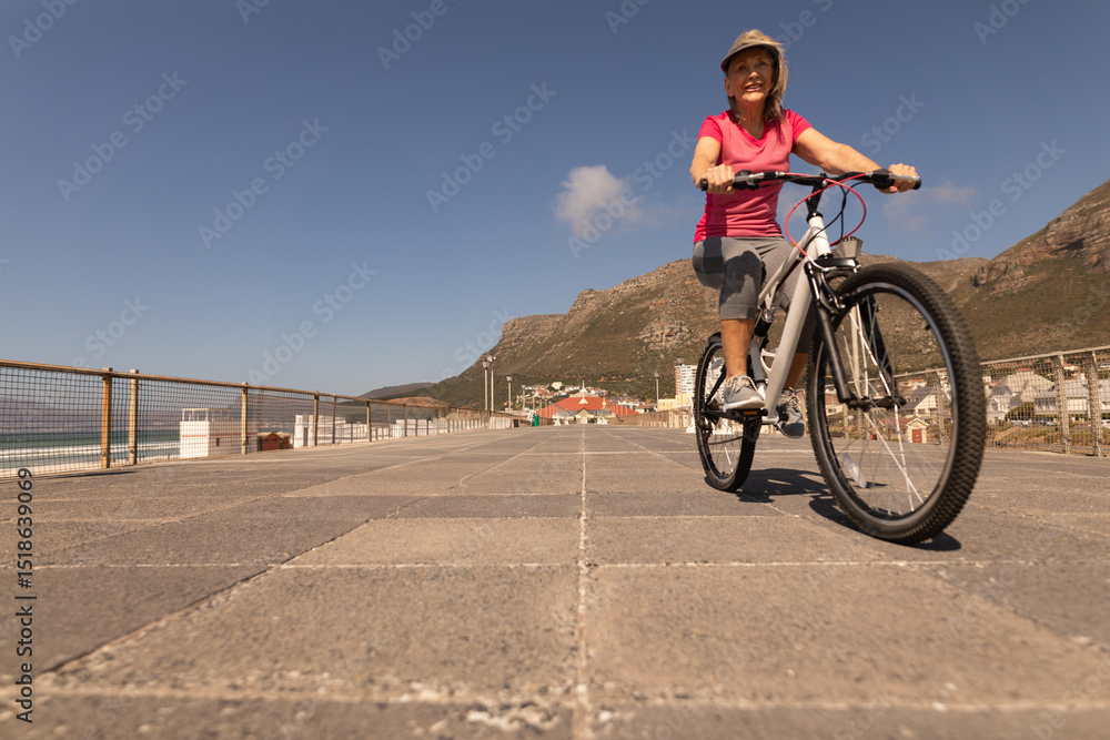 Obraz premium Senior woman wearing sunhat riding white mountain bike on coastal promenade by railing, copy space
