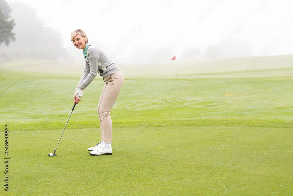 Fototapeta premium Middle-aged woman in golf gear gripping putter lining up golf ball on putting green with flagstick