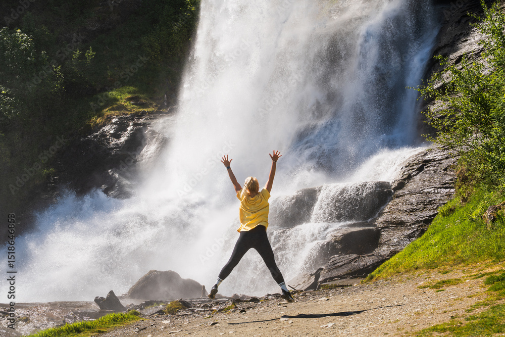 Obraz premium Joyful traveler jumps with delight against the backdrop of a powerful waterfall, enjoying freedom and the beauty of Norway's wild nature.