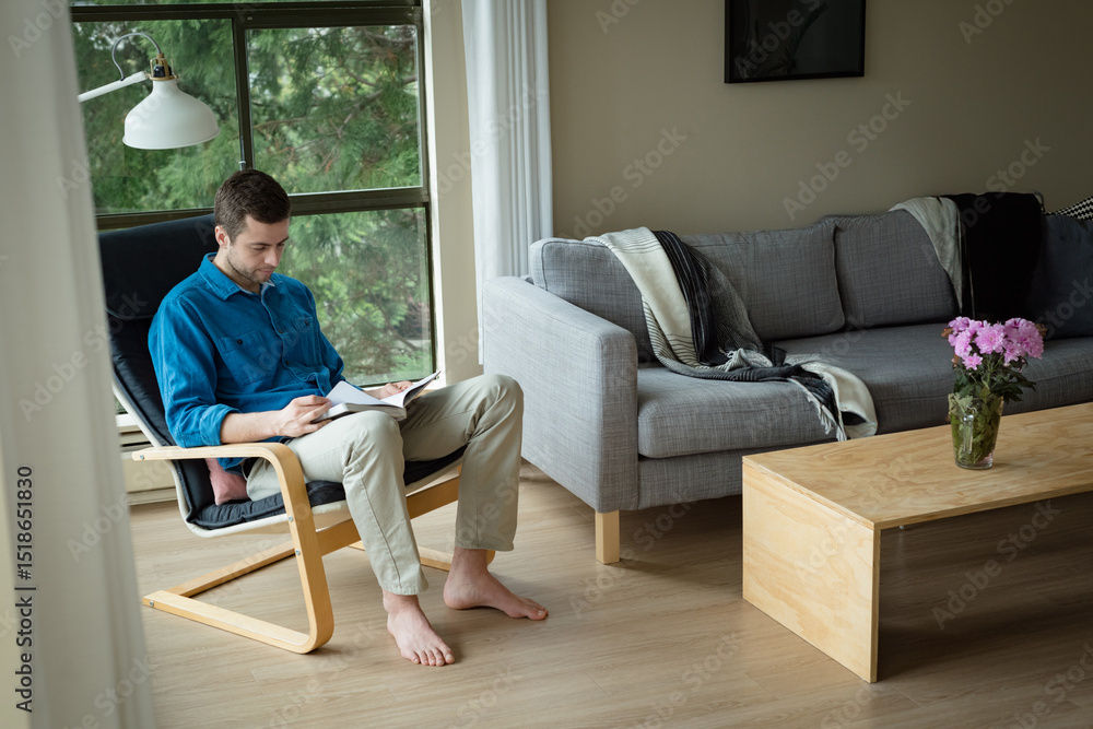 Man in 30s sitting on chair in living room reading document holding pen, copy space Man in 30s sitting on chair in living room reading document holding pen, copy space
