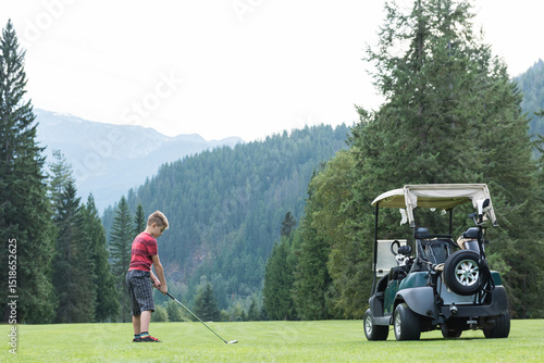 Boy aged eight holding golf club and ball on mountain fairway beside parked cart, copy space