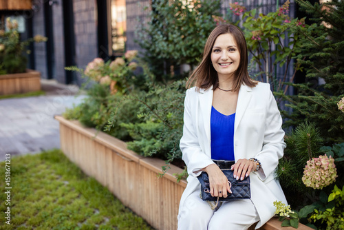 Young cute woman sitting on park bench in modern city, smiling and looking at camera