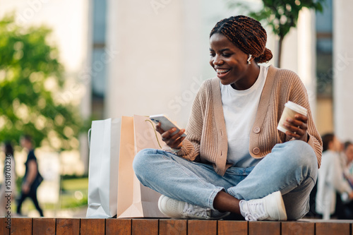 Konstfotografi Smiling shopper using smartphone and holding coffee with shopping bags