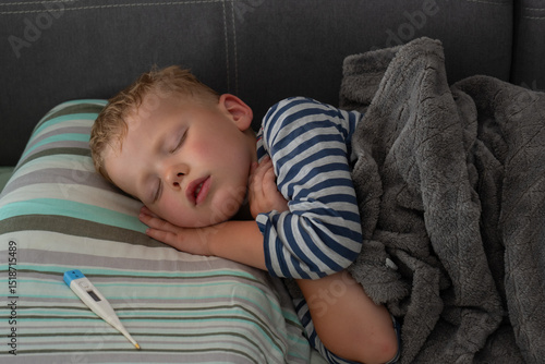 Young child sleeping under gray blanket with a digital thermometer nearby on a striped pillow. Concept of home healthcare, illness recovery, and parental care.