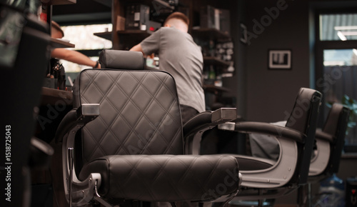Tablou pe pânză Black leather barber chairs line a modern barbershop station, as a barber in the background prepares tools on a wooden counter, highlighting sleek design, comfort, and professional ambiance