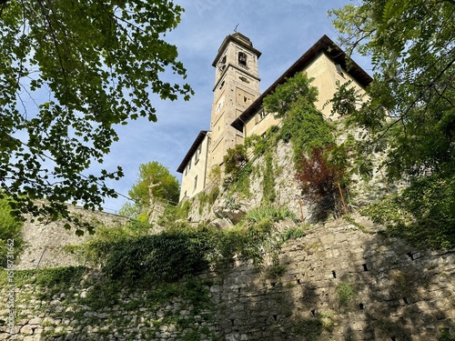View from below of the Sanctuary of Madonna del Soccorso in Ossuccio, Lake Como, Italy, with stone walls, bell tower and lush greenery under a bright blue spring sky