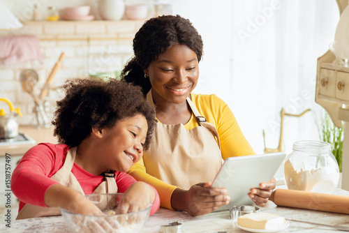 Fototapet Happy black mom and her little daughter checking dough recipe online on digital