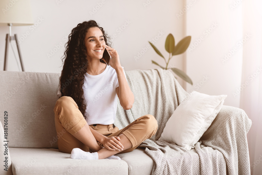 Happy brunette woman with long curly hair having conversation on mobile phone while sitting on couch in living room and looking at copy space. Young lady talking on phone and smiling Happy brunette woman with long curly hair having conversation on mobile phone while sitting on couch in living room and looking at copy space. Young lady talking on phone and smiling
