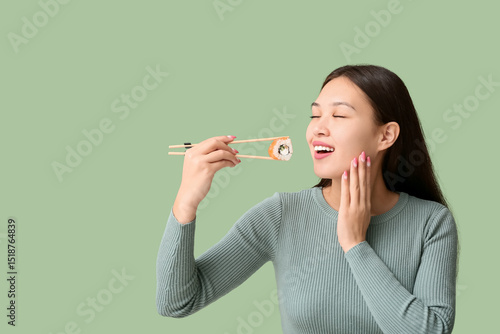 Obraz na plátně Beautiful young Asian woman holding tasty sushi roll with chopsticks on green ba