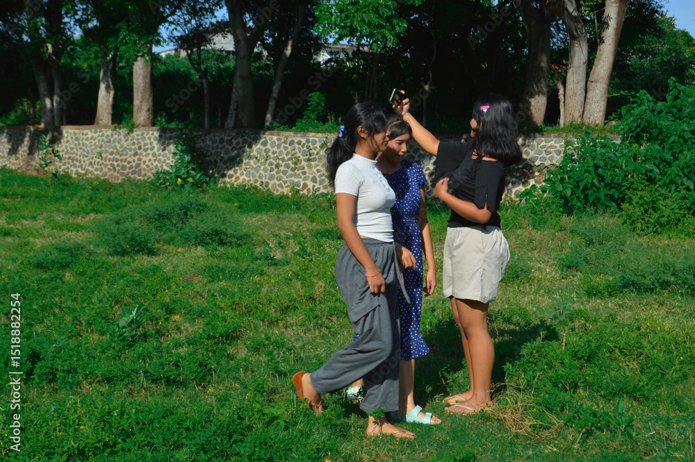Fototapeta premium One girl holds up her phone to take a selfie while the other two walk toward her across the grassy field.