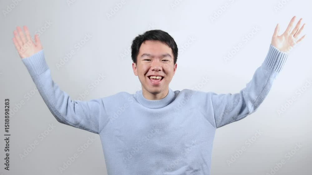 A young Asian man in a light blue sweater shakes his arms enthusiastically in a joyful celebratory gesture. The scene conveys excitement, happiness, or a casual cheer in a studio setting. A young Asian man in a light blue sweater shakes his arms enthusiastically in a joyful celebratory gesture. The scene conveys excitement, happiness, or a casual cheer in a studio setting.