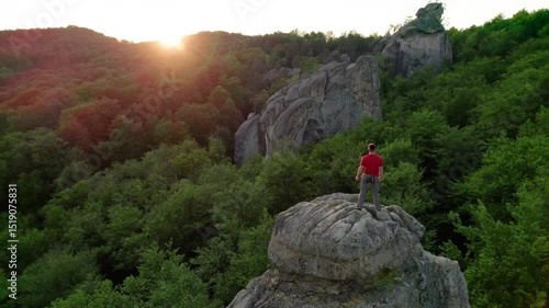 Aerial view of climber standing atop rock formation at Dovbush Rocks in Carpathian mountains, Ukraine. Sun sets, casting warm glow over lush, green forest, distant hills, creating breathtaking view.
