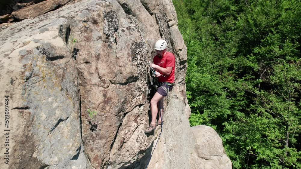 Aerial view of male rock climber ascending rugged limestone cliff with harness and rope for safety. Sportsman climbing on vertical large boulder at Dobvush Rocks in Carpathian mountains, Ukraine. Aerial view of male rock climber ascending rugged limestone cliff with harness and rope for safety. Sportsman climbing on vertical large boulder at Dobvush Rocks in Carpathian mountains, Ukraine.