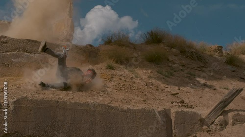 Man with toy gun stumbling  down earth mound as explosion goes off in super slow motion