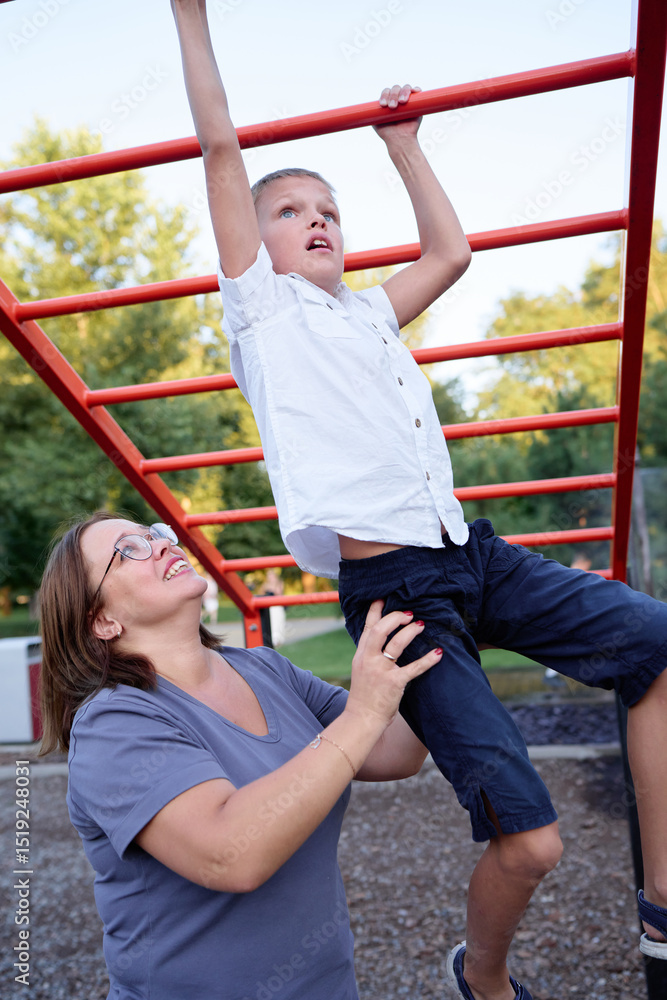 Obraz premium Woman assists her son as he attempts crossing red monkey bars in a park. A motivational moment of compassion and learning, capturing essence of support and encouragement in family activities outdoors.